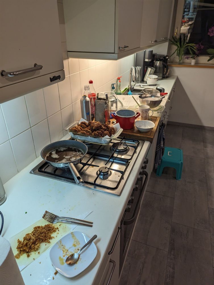 Küche nach dem Kochen von Kaspressknödl und Thai-Curry-Pfanne. Von vorne nach hinten: Teller mit Curry Pasten Reste und Kaffeelöffel. Küchenpapier mit ca 4 EL frittierten Käseresten. Pfanne mit 1 Tasse gebrauchten Butterschmalz. 19 Kaspressknödl. Brett mit zusammengestellten schmutzigen Küchen Utensilien. Doppelspüle voll mit schmutzigem Geschirr. SodaStream, Wasserkocher, Kaffeevollautomat