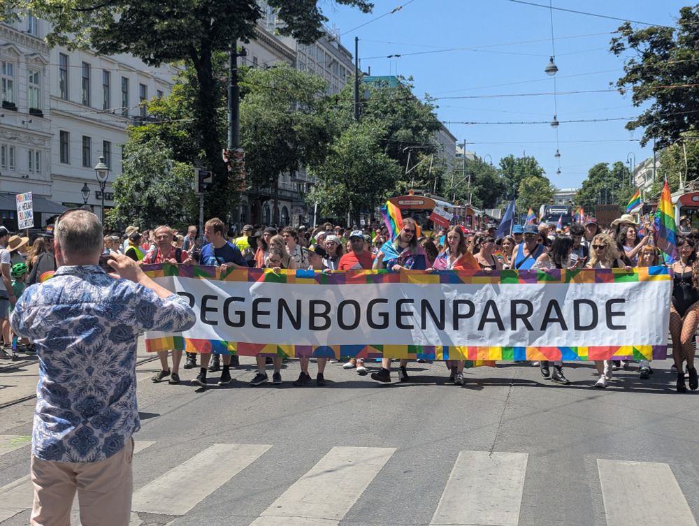 Anfang Regenbogenparade 🌈 aka Vienna Pride ca. Höhe Karlsplatz. Großes Transparent mit der Aufschrift: Regenbogenparade, dahinter viele bunte Menschen und die Techno Trams der Wiener Linien.