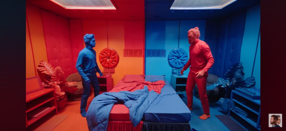 A screenshot of Rhett and Link’s “We Spent 24 Hours Locked in Red and Blue Rooms”. The room is painted fully red on our left and blue on our right. Link stands on the left painted blue and Rhett is opposite of him painted red. A bed is between them, split but the respective rooms color. 