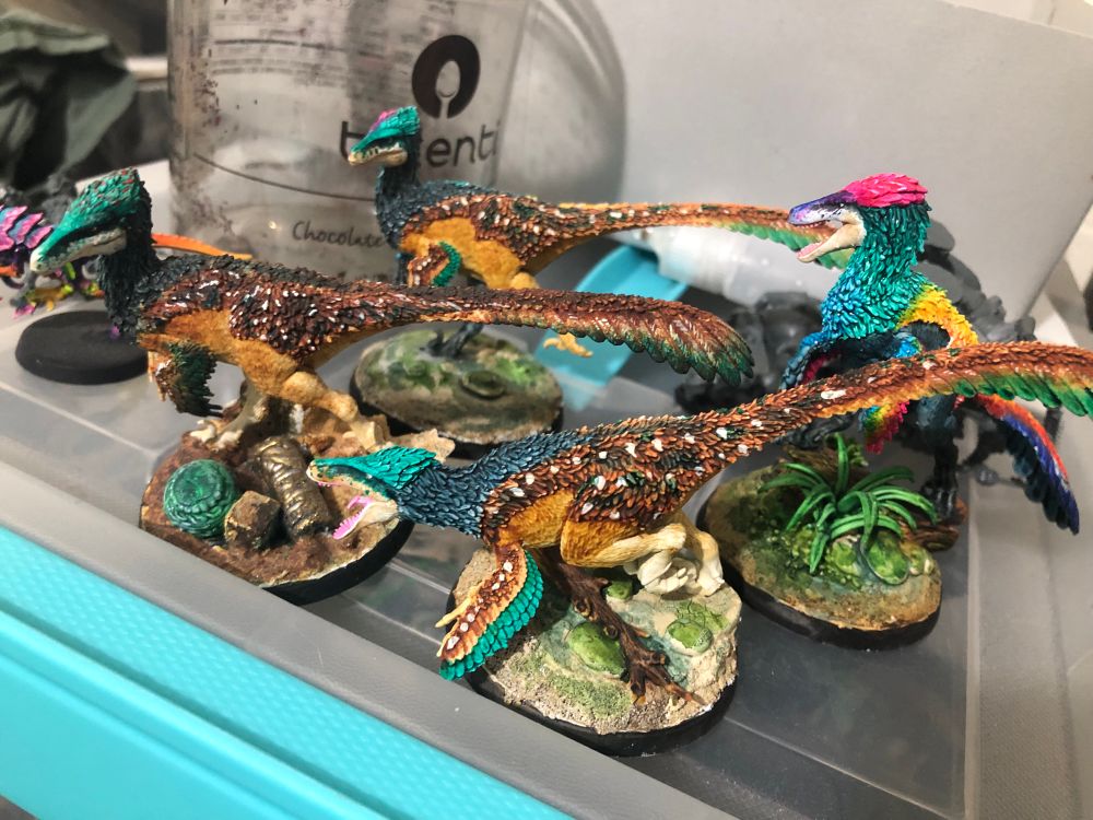 Four miniature Utahraptors; four of them are painted with dappled brown and white with green crests and wing feathers, one is yellow, pink, and green. They are in various poses.