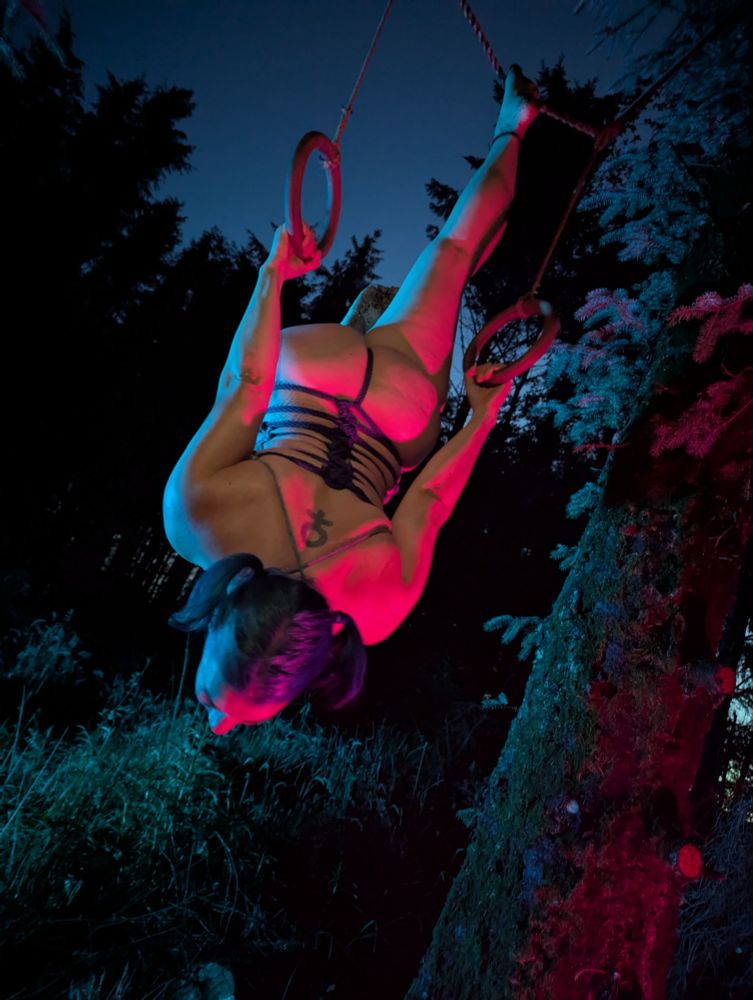A person hanging inverted from gymnastic rings in a pine forest, wearing a rope corset, shot from the back in bisexual lighting