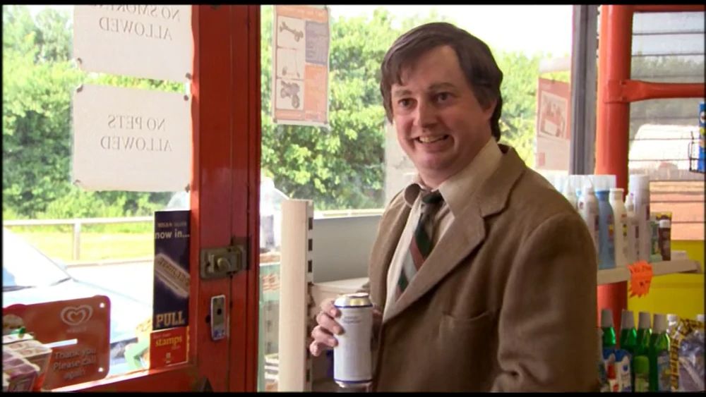 A British man (played by comedian David Mitchell) in a frumpy suit stands at the counter of a corner shop. He has a gormless smile and is holding a tall can of lager beer. 
From the "That Mitchell and Webb Look" sketch "Corner Shop."
