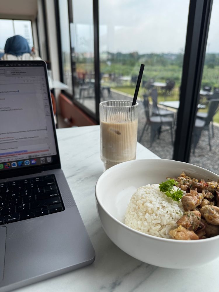 Photo of spicy chicken matah rice and ice coffee beside the laptop. and also beside of the table is big glass window showing farm