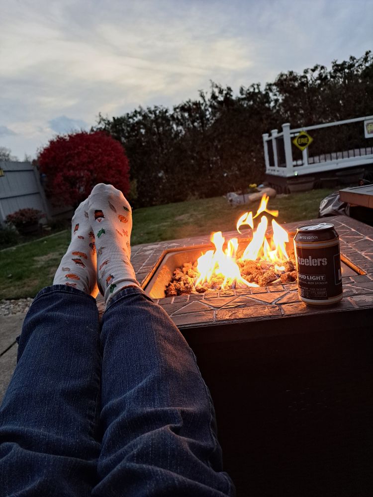 Socked feet next to the flames of a propane fire table, with a can of Bud Light Steelers beer can, with a yard in the background that has the garden cleared out.