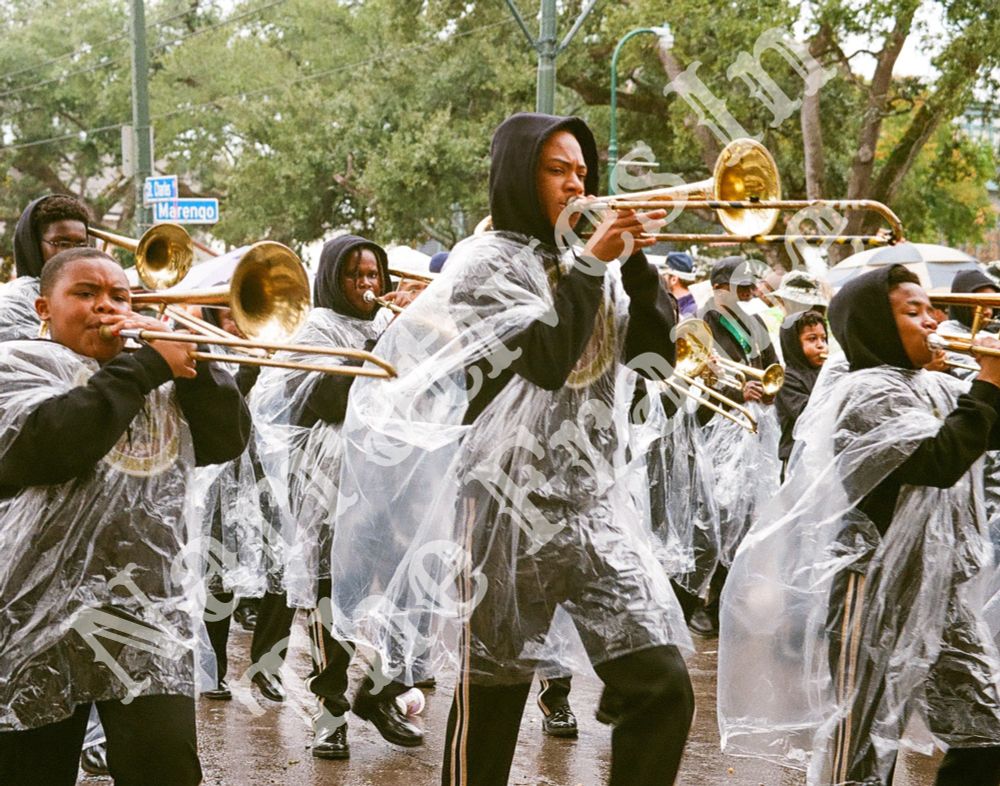 A group of Black middle and high school boys marching in a Mardi Gras parade, playing brass instruments. Due to earlier rain, they are wearing clear plastic ponchos with black tracksuits visible underneath. Trees line the background, and a small blue cross-street sign is visible in the distance.