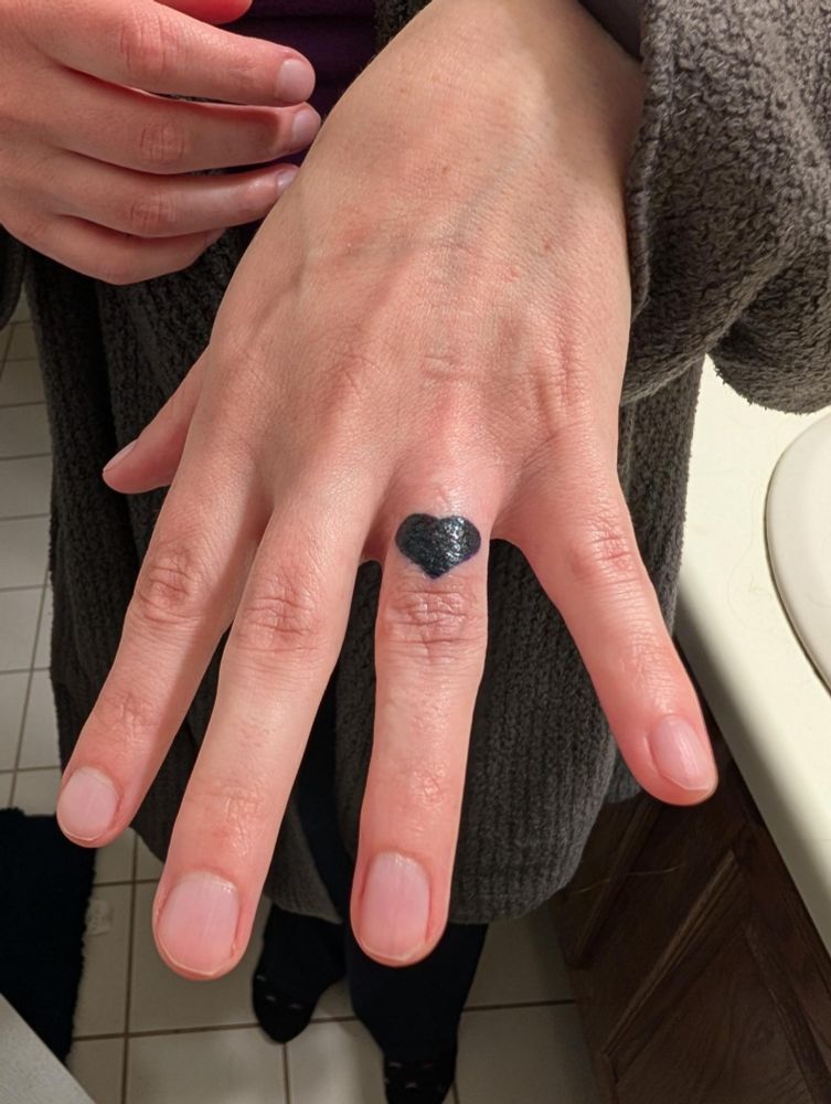 A picture of my hand, with my new tattoo! It's a black heart on my ring finger, right where a ring would sit. I'm white, and my nails are short and unpainted. 