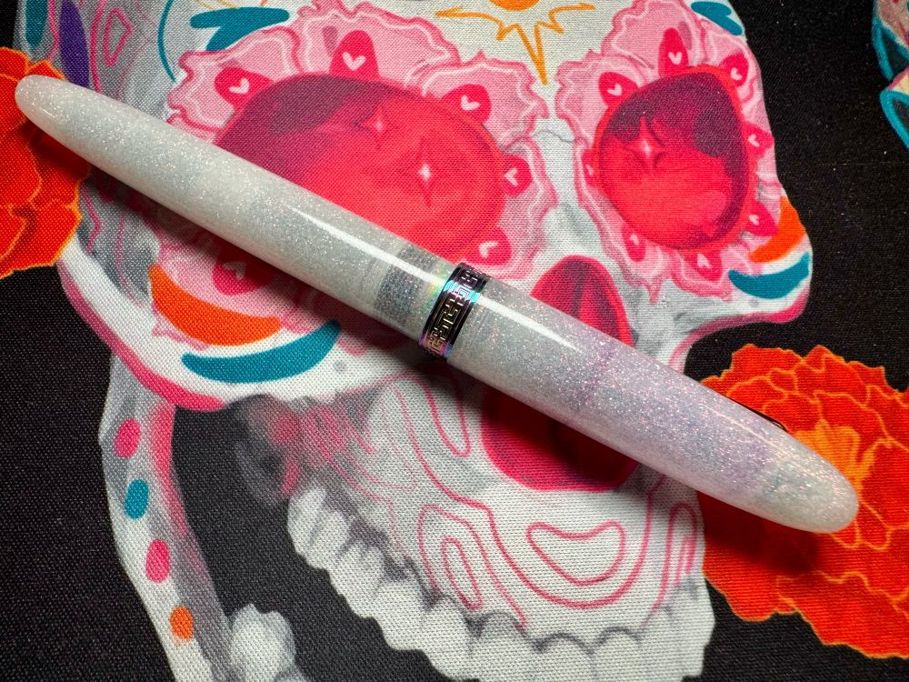 Closeup of a Navahlur Key West Pride 2024 cap with staining. The pen is closed and white with sparkling glitter. The background is a desk mat with a colorful pink and teal sugar skull. 