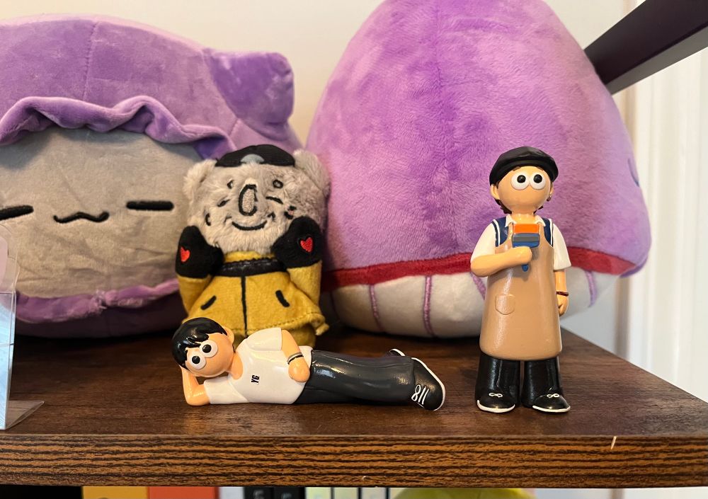 A wood shelf with a squishy Suga Cat plush wearing a purple hoodie, a purple BTS whale plush, a mini Namjoon koala plush wearing his outfit from the BTS Go, Go heart performance at MBC in 2017, 3D figurine of Yoongi lying down wearing a white FG shirt and black pants, and 3D figuring of Yoongi from his painting live where he’s wearing a sweater vest and apron. 
