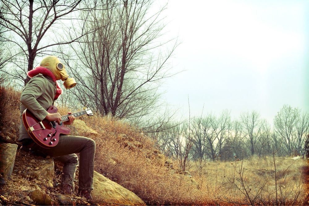 A man in a golden gas mask playing a red guitar in a barren meadow.