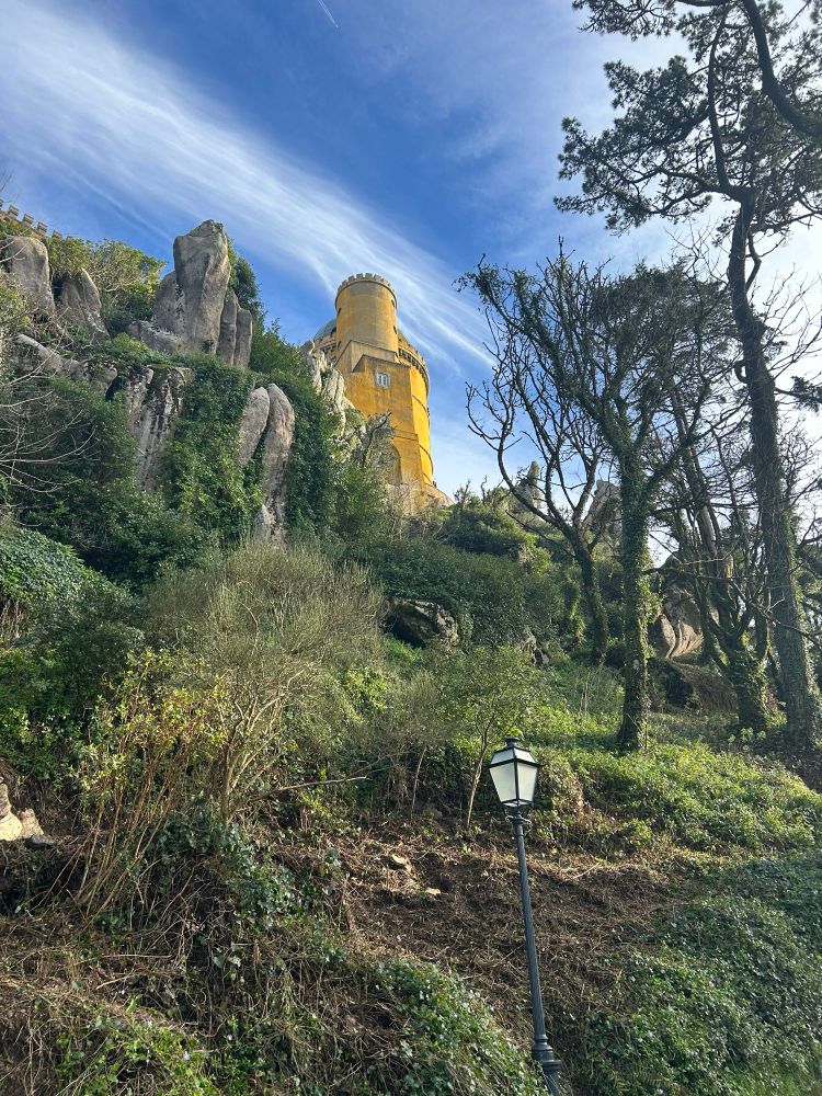 A yellow cylindrical tower, part of Palácio da Pena, rises above a rocky, green hillside with a lamppost and trees under a blue sky with wispy clouds.
