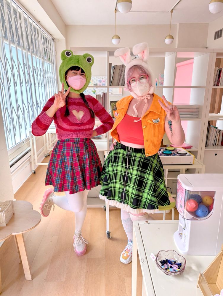 Me and @sailorhg.com cosplaying DDR characters Kero-Zukin and Oshare-Zukin. Kero-Zukin has a green frog hood and red plaid skirt and Oshare-Zukin has a bunny hood and green plaid skirt. 