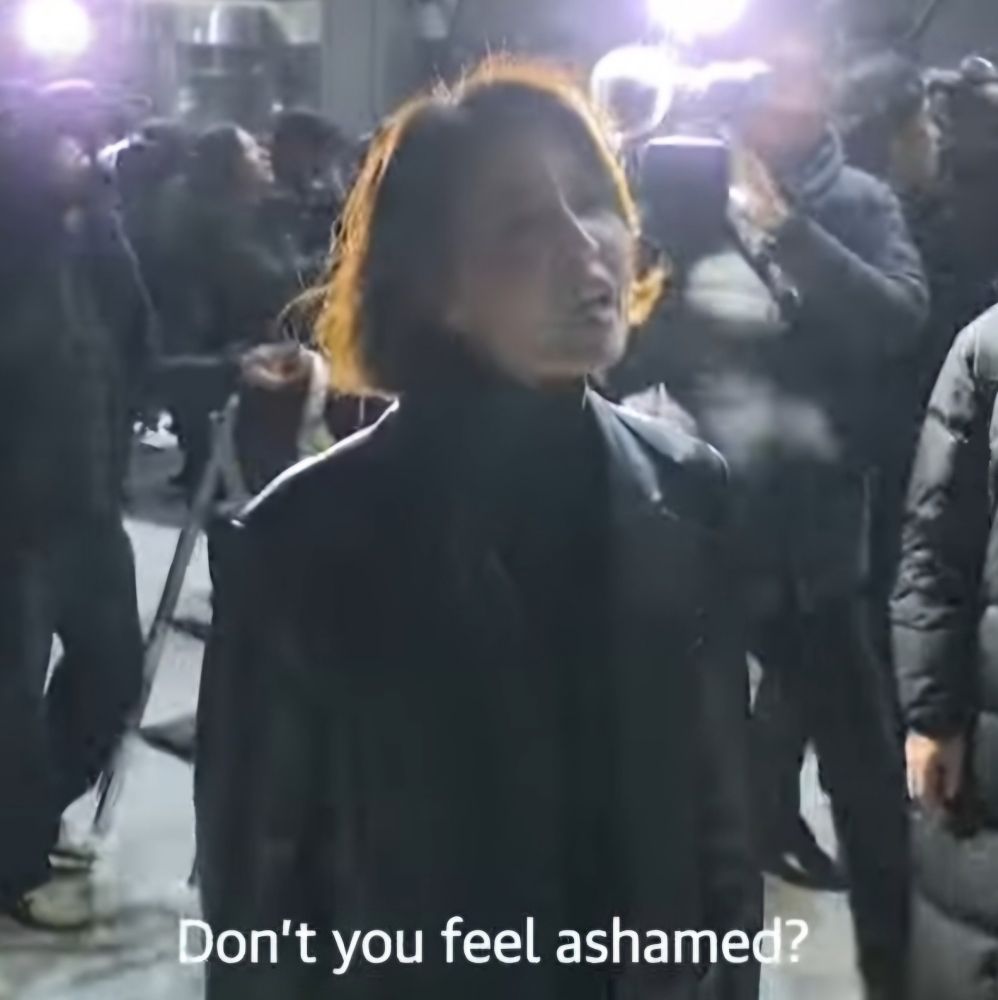 Woman standing in chaotic crowd shouting "Don't you feel ashamed?"