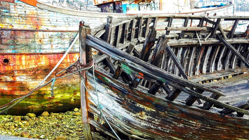Cimetière de bateaux de Camaret.
Les bateaux sont là depuis des années. Au premier plan, un bateau en bois bien abîmé. Derrière,  on aperçoit un autre bateau coloré de vert par les algues et d'orange par la rouille..ils reposent sur un lit de galets.