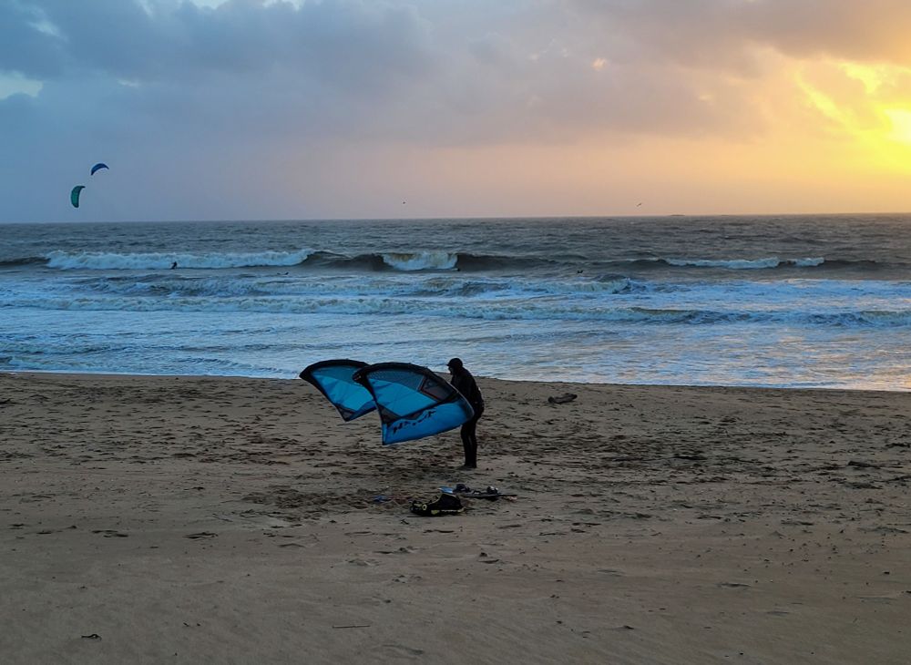 Un surfer replié sa voile sur le bord d'une plage, au soleil couchant