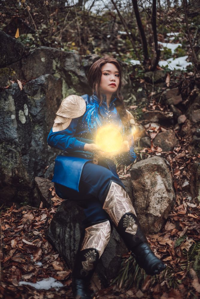 me as talasyn of sardovia, sitting on a rock wearing a blue leather tunic and blue trousers, gold pauldrons and gold embellished black boots. hands are in front cradling a ball of light 
