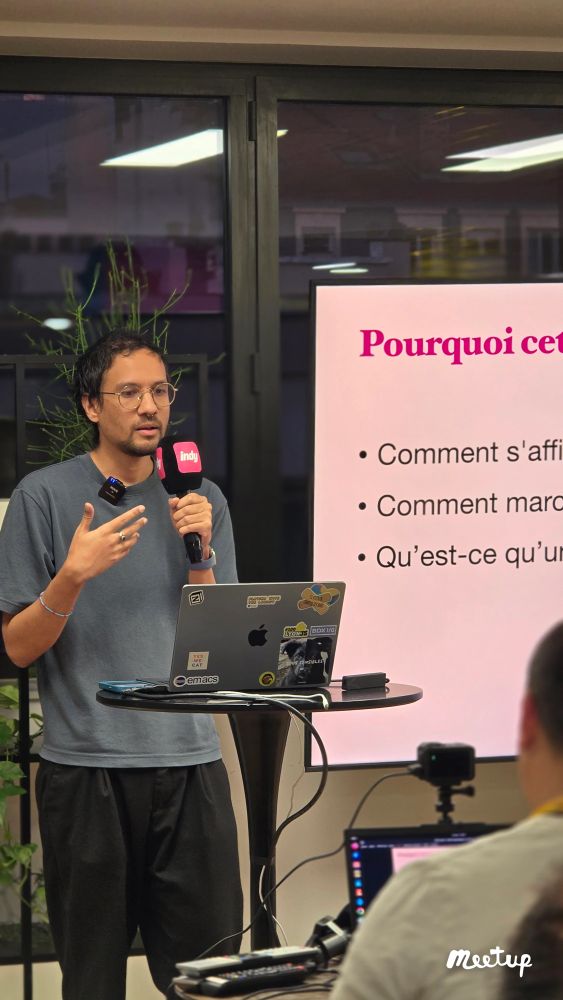 Je m’exprime à une conférence et je parle de frameworks JavaScript. Je parle dans un micro rosé que je tiens, on voit mon macbookpro couvert de stickers et on voit un écran de présentation en arrière plan