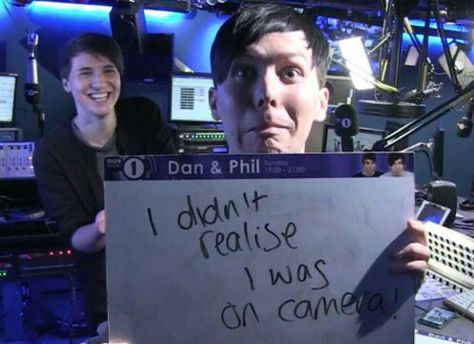 Dan and Phil at the BBC Radio Station. Phil holds up a small whiteboard that reads "I didn't realize I was on camera!" while Dan laughs in the background.