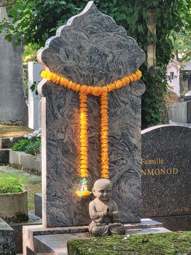 Eine Kette aus orangen Tagetes zieren einer oriantalisch geformten Grabstein. Davor sitzt ein betender Buddha 