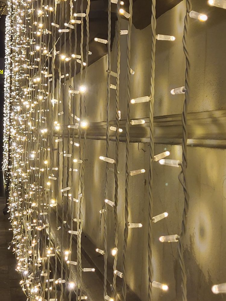 An einer weißen Gebäudefasse sieht man große Streifen einer weißen Lichterkette, die einen Vorhang bildet.