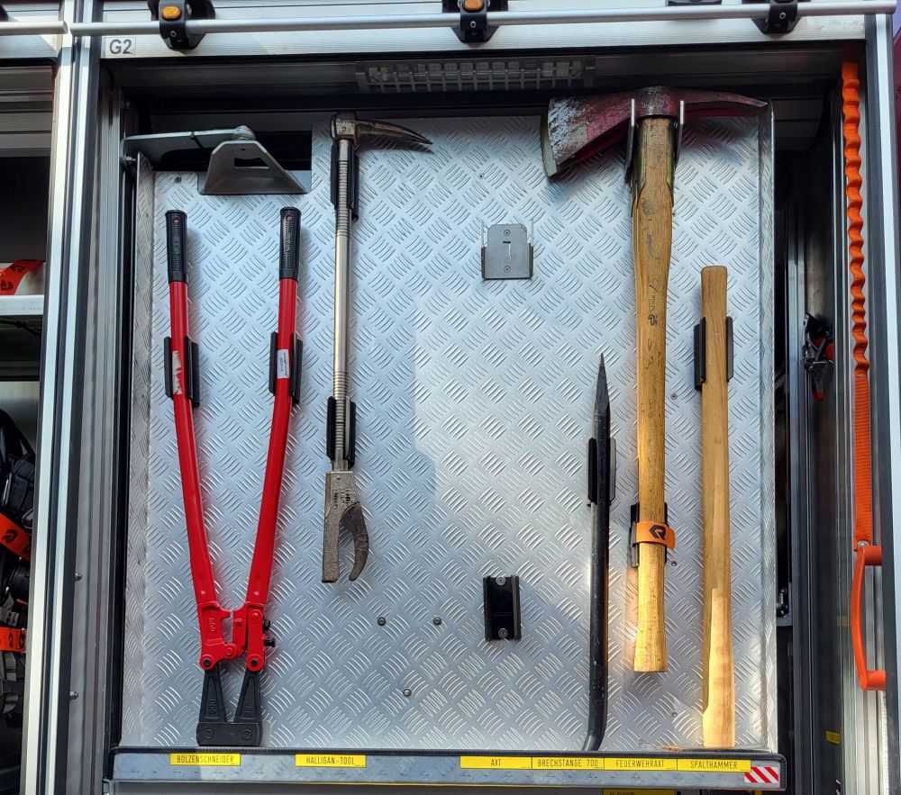 Eine große Zange, ein Greifer und zwei Äxte (teilweise zerlegt) sind an einem Metallgitter aufgehangen.