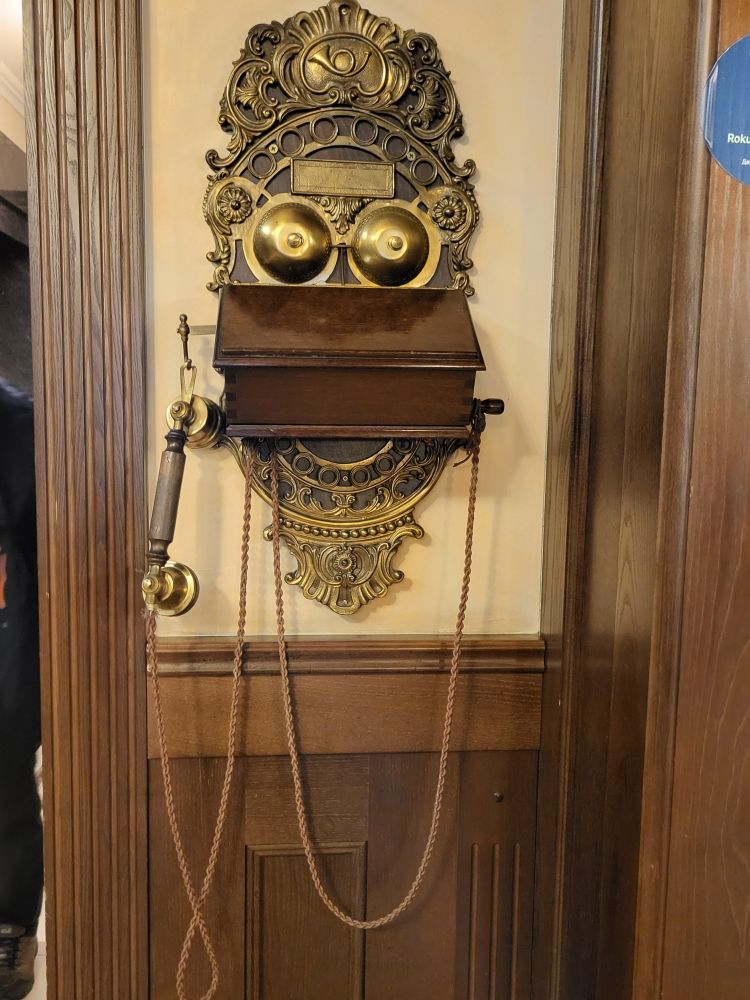 An der Wand ist ein sehr altes Telefon. Der Hörer hat eine sehr lange Schnur. Gold/ Bronzefarbig die Verzierungen. Mittig ein Holzkästchen.