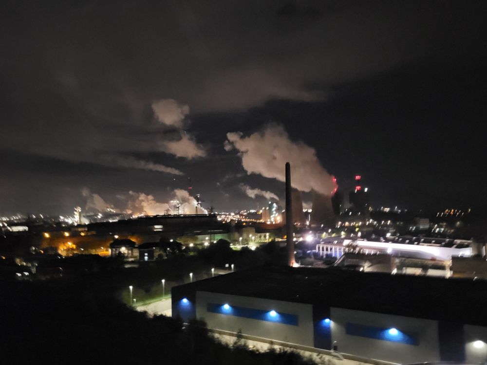 Zu sehen ist eine Duisburger Industrielandschaft bei Nacht. Im Vordergrund eine Halle beleuchtet von 5 blauen Lampen. Dahinter ragt mittig ein Kaminschlot. Dahinter zwei große Kühltürme die Dampf ablassen. Irgendwelche Türme mit roten Warnlichtern Dahinter. Mittig bis zum linken Seitenrand sieht man viel Dampf unter nicht mehr erkennbaren Industriengebäuden.