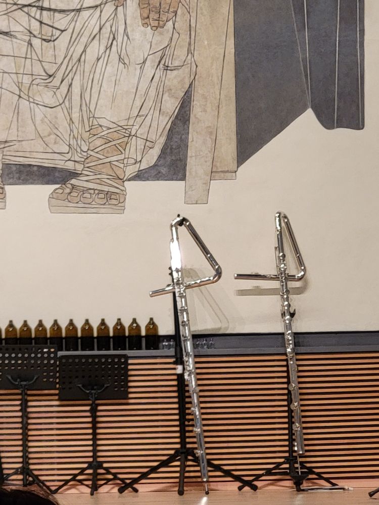 Zwei Bassquerflöten lehen in Ständern. Sie haben quasi ein Dreieck im Instrumentenverlauf. Das erzrugt tiefe Töne. Links Apothekerflaschen und Notenständer.