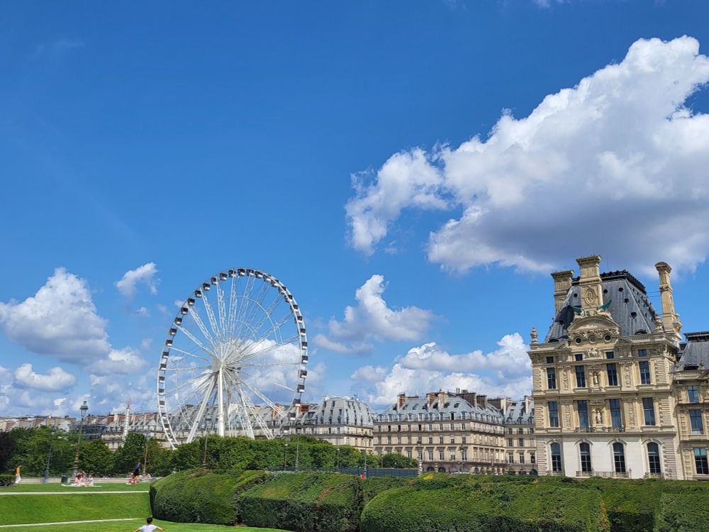 Vor einem spektakulären blauen Himmel mit einige weißen Wölkchen sieht man dasGrün eines Parks. Im Hintergrund ein Riesenrad und dahinter prächtige Gebäude bis zum Horizont.