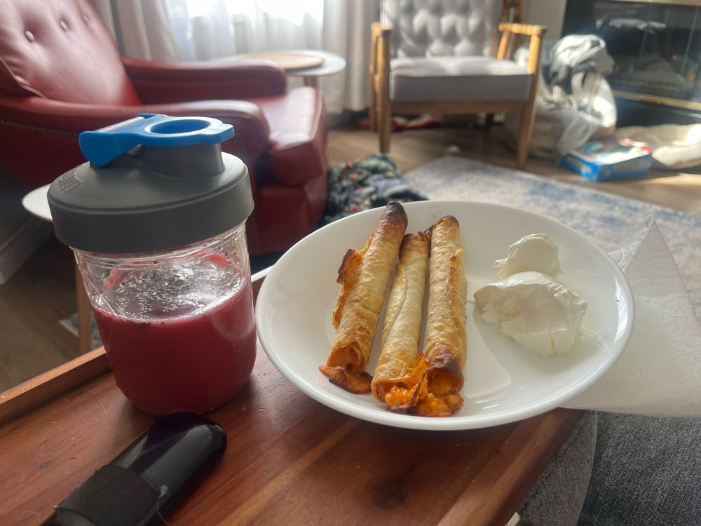Taquitos in a bowl with generous helping of sour cream. A glass of ginger beet juice with chia. Messy living room in background with sunlight coming through a window. 