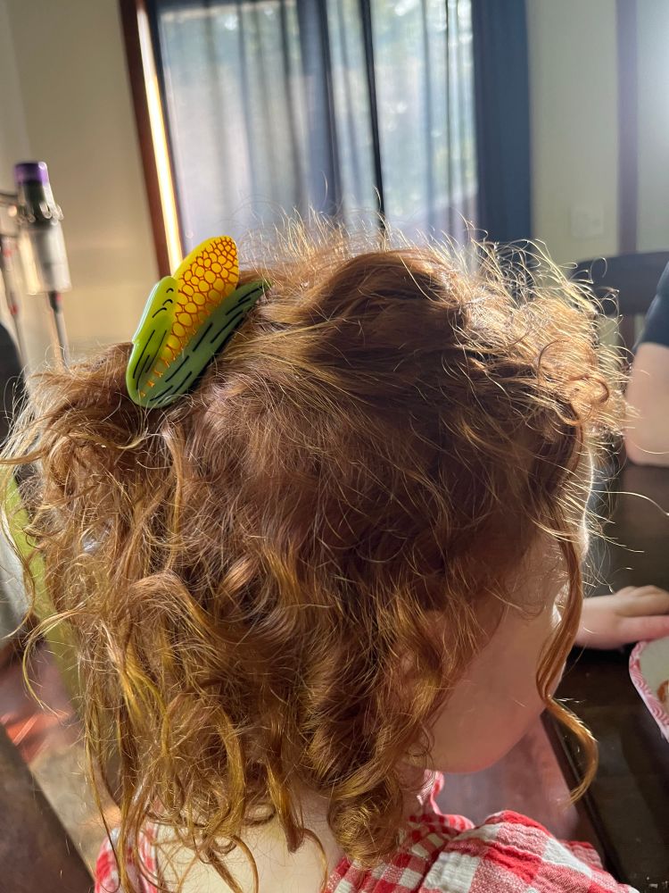 Side picture of a small girl with curly red hair.  The top and sides of her hair are pulled back with a clip that looks like an ear of corn.