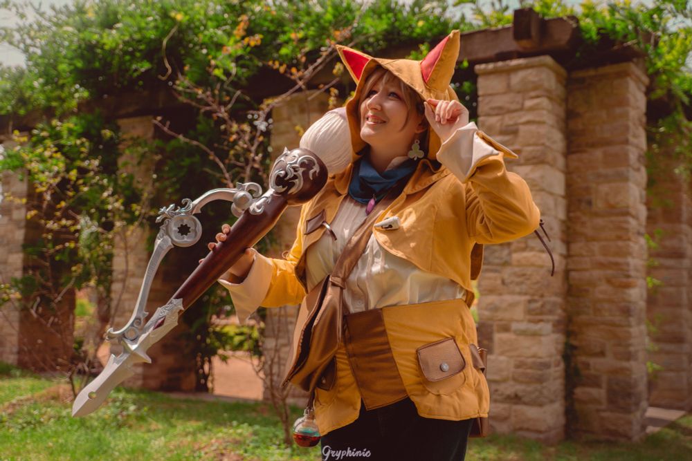 A photo of a cosplayer dressed up as Pictomancer Krile from final fantasy xiv. Some stone pillars and greenery are in the background.