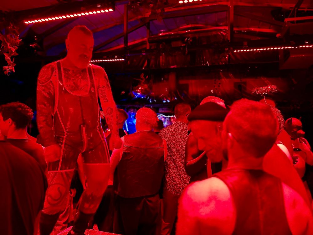 Tattooed dad in a latex singlet dances on the block on a crowded dance floor. 