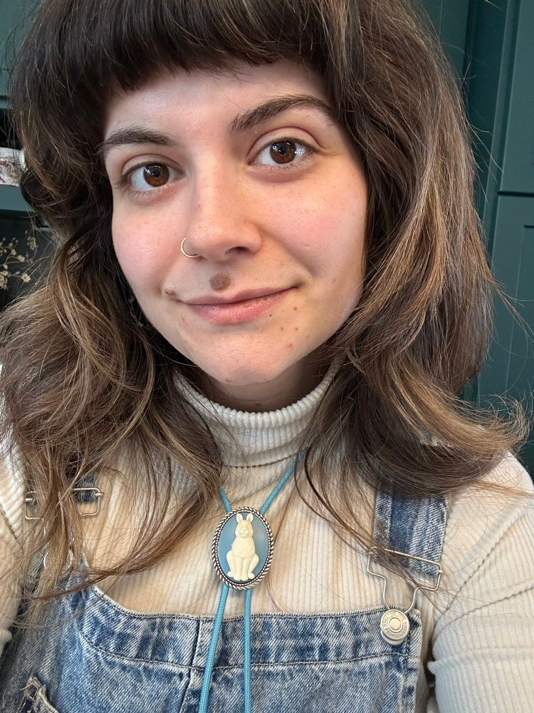 A white cis Bosnian woman with long wavy brown hair is smiling at the camera. She wears a cream turtle neck and a blue bolo tie with a white sculpted bunny as its middle. 