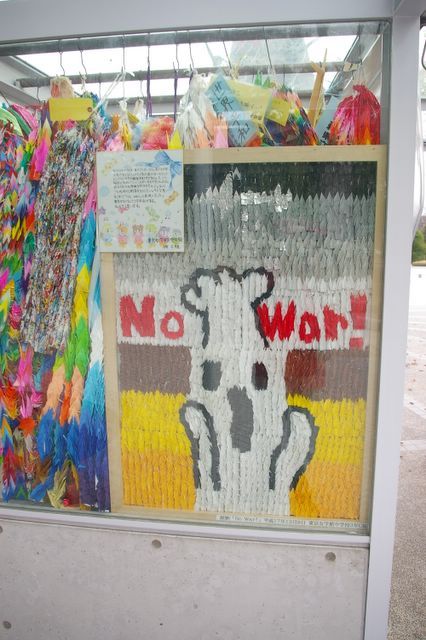a cabinet containing origami cranes and a screaming figure saying "No War!" made from coloured origami cranes.