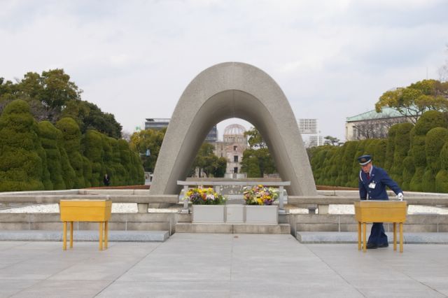A concrete arch mid frame. In front two wooden tables, one being adjusted by a man in a blue uniform with cap. Trees in the background, through the arch a ruined building with the skeleton of a dome on top can be seen.