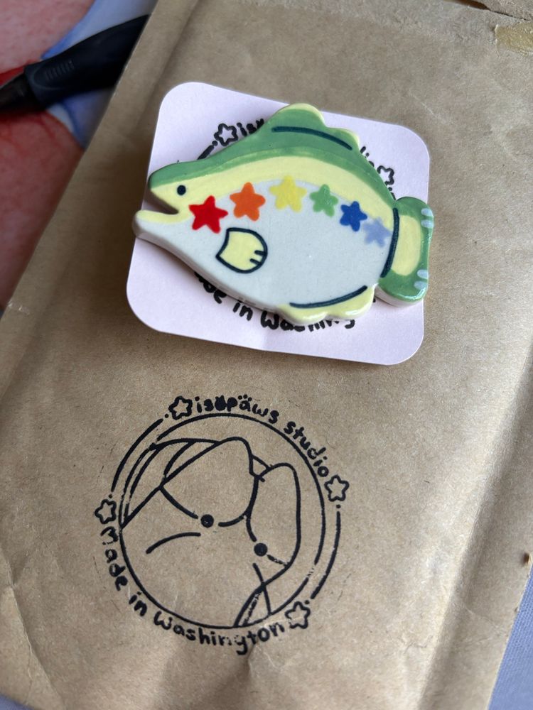 A rainbow trout pin with rainbow stars on it from the ceramicists Isopaws 