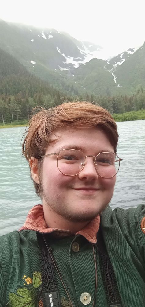 me! guy with short light brown/orange hair with a short beard and glasses standing in front of a lake and a glacier, smiling wearinga  green jacket