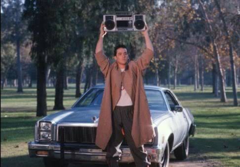 Boombox scene from Say Anything. Lloyd, a young white manis dressed in a brown coat and dark pants. He’s standing in front of a (now) vintage car. I don’t know what type, sorry. It’s white or silver. He holds a boombox above his head. 