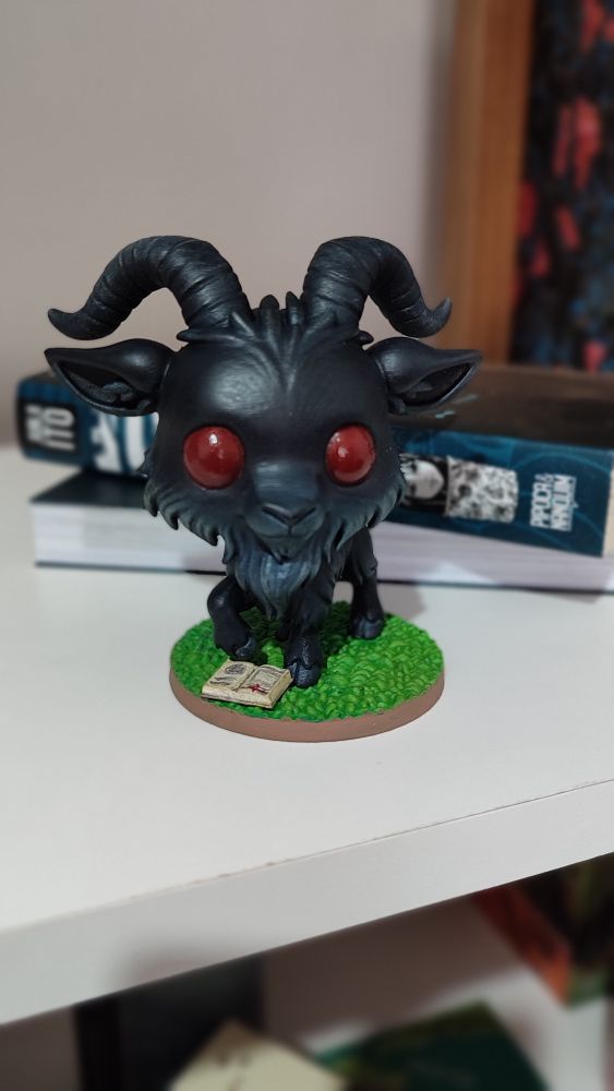 Imagem mostrando uma miniatura personalizada de Black Phillip, um bote todo negro que seria a manifestação do Diabo no filme A Bruxa. No centro da foto temos a imagem do bode negro com olhos vermelhos e estilização de modo que lembra o estilo chibi. A mesma está posicionada em uma prateleira de livros com o segundo volume do mangá Tomie ao fundo 