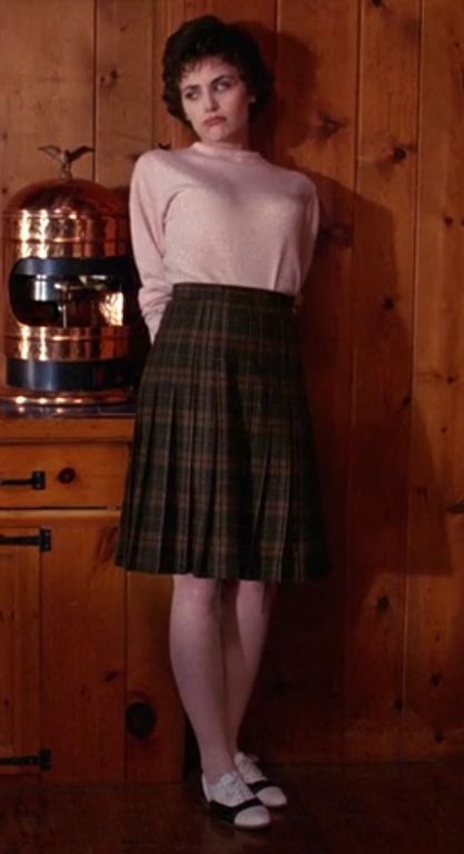 A picture of Donna from Twin Peaks. She is dressed in a pink top with a plaid skirt. She is leaning against a wooden paneled wall with her arms held behind her, as she looks off to the side with a frustrated, bored, pained expression. 
