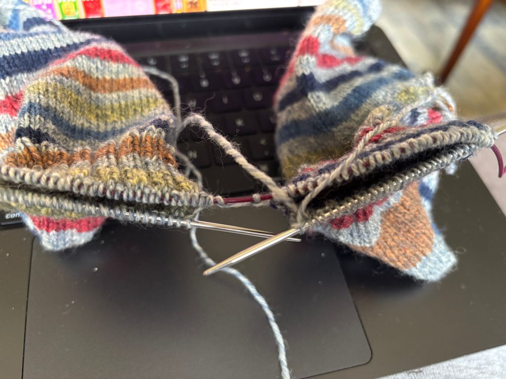 A pair of striped socks with reversed stripe patterns of dark blue, medium blue, yellow, orange, and red with gray stripes between as a background on a single circular needle sitting on the keyboard of an open black laptop. The sock on the right has two working yarn tails in and near the last worked stitch because the yarn for the left sock was not dropped to pick up the correct yarn for the right sock