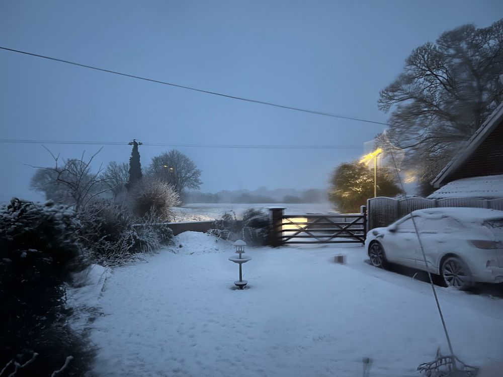 Photograph of a front garden, drive and car, and fields and trees beyond all covered in white snow. 