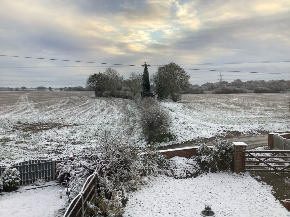 Photograph of fields with a hedgerow separating them with a lane and two garden lawns in the foreground of the photo. Everything is covered with a light dusting of snow. The sun is breaking through the grey clouds. 