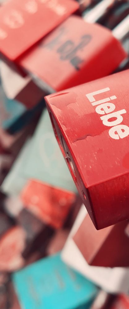 Fotoausschnitt von Liebesschlössern. Ein Rotes bestimmt mit dem Wort "Liebe" (in weißen Buchstaben) das Bild, es ist nur halb zu sehen. Das Schloss ist noch feucht vom Regen. Die anderen Schlösser (rot und blau) sind nur unscharf zu erkennen.