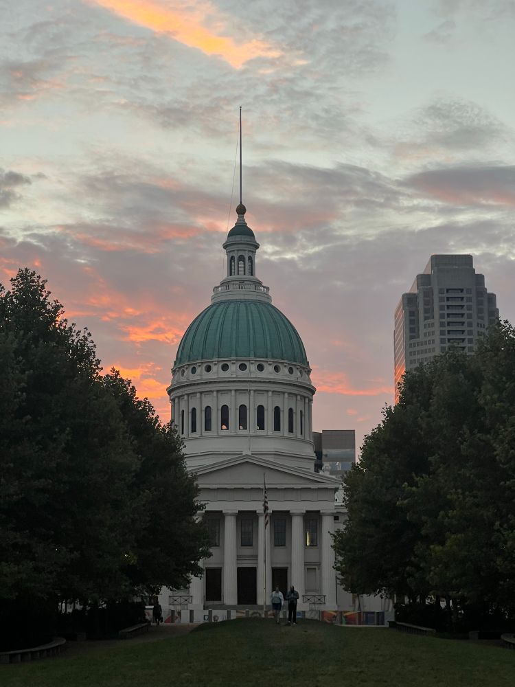 Sunset sky in the background of a courthouse with white columns and a green domed roof. 