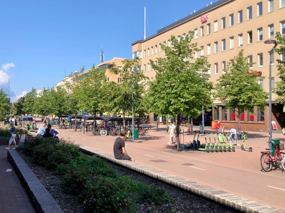 The dedicated two cycling lanes and bike parking with street trees and places for folks to site down in Joensuu, North Karelia. An SUV is stationary after driving on the cycling lane on the left.