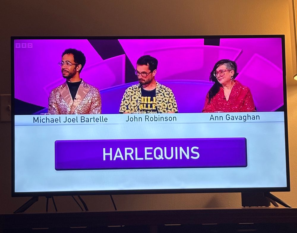 Photo of a TV with a quiz team named the Harlequins made up of three people. On the left a man is wearing round, colorful glasses, a black t-shirt, and a sparkly pink blazer made of satin and sequins. Next to him another man is wearing a t-shirt with yellow letters under a sunflower patterned short sleeve button down shirt. On the right is a woman with long hair and long dangly earrings. One side of her head is shaved and she is wearing a pink sequined blazer. 