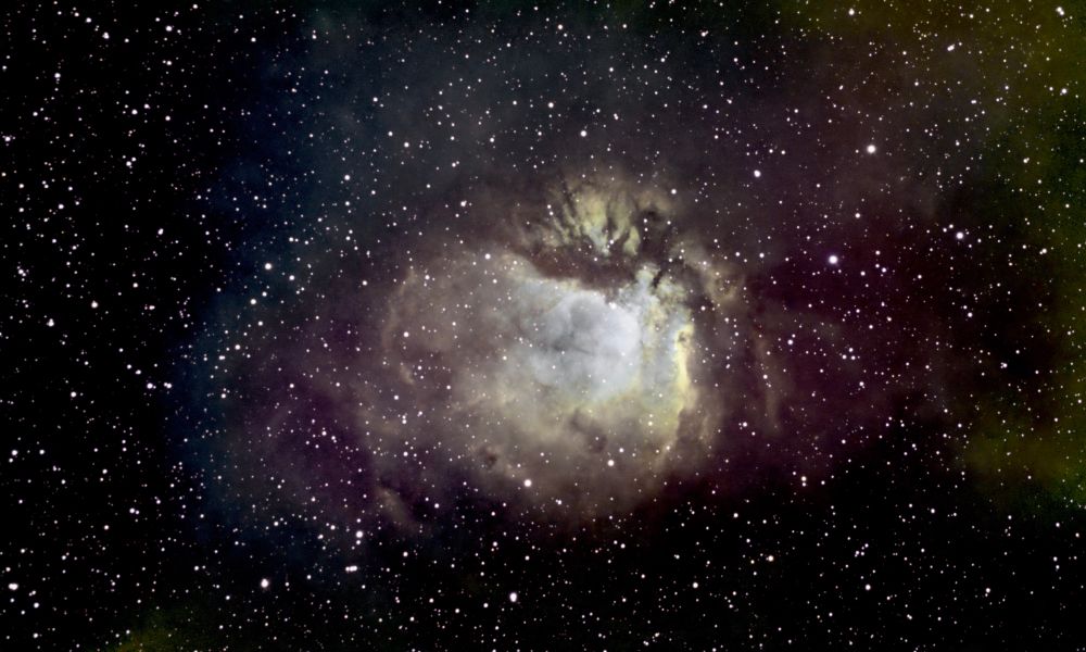 A bright central circular image in pale blues and greens surrounded by red toned nebulosity and stars. 