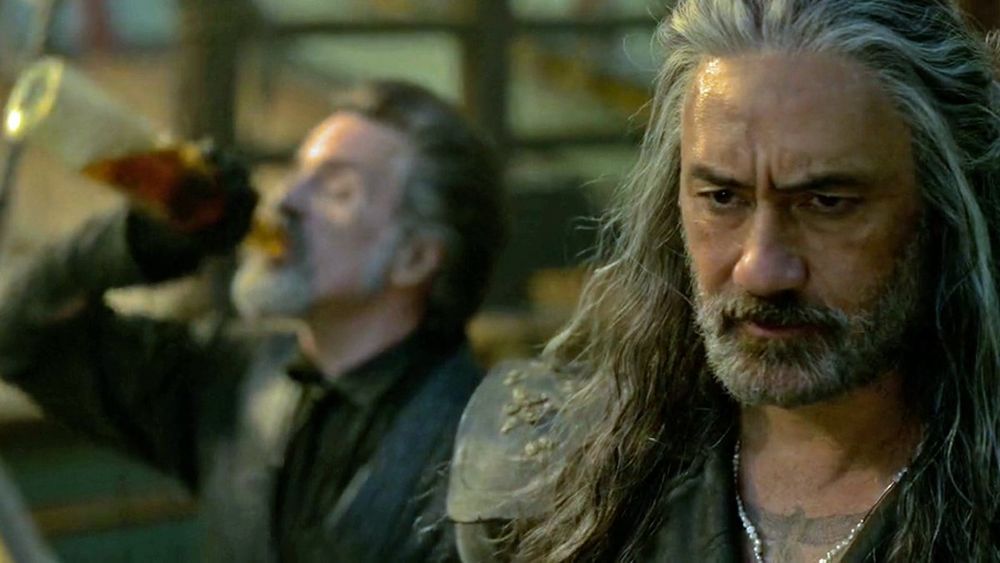 Ed Teach, a pirate with a short gray beard and long hair, wearing leathers and looking pensive. Behind him, a blurry Izzy Hands, also a pirate in leathers with a short gray beard but with shorter hair, swigs heavily from a bottle of rum.