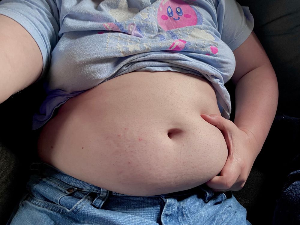 A photograph of my own gut, exposed by my t-shirt being pulled up. I lightly grab at it with one hand while taking the photograph with the other.